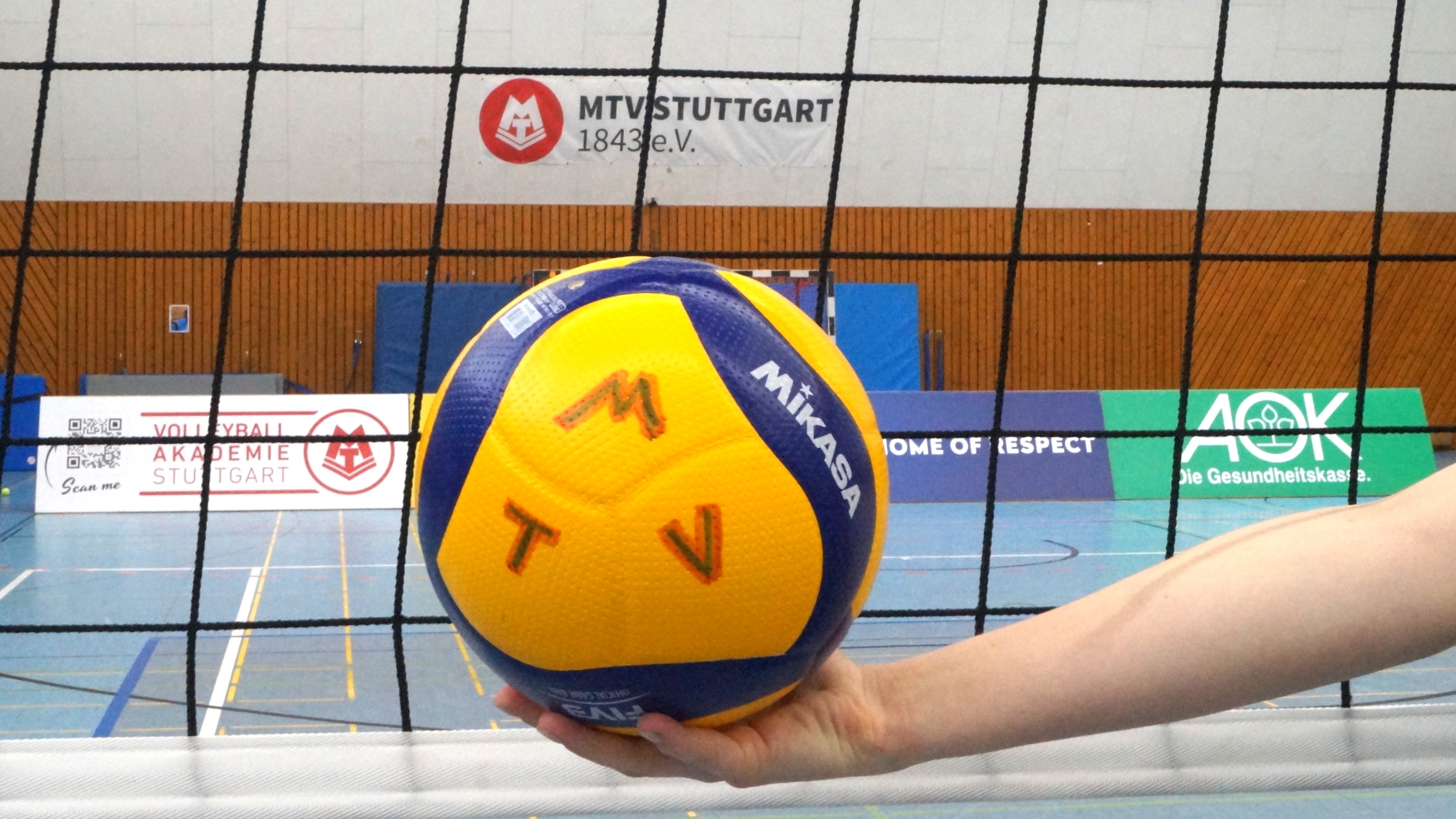 Bild zeigt einen Volleyball, der von einer Hand vor einem gespannten Volleyballnetz gehalten wird. Der Ball trägt die Bezeichnung "M T V"; im Hintergrund ist der Schriftzug "MTV Stuttgart 1843 e.V." zu sehen.